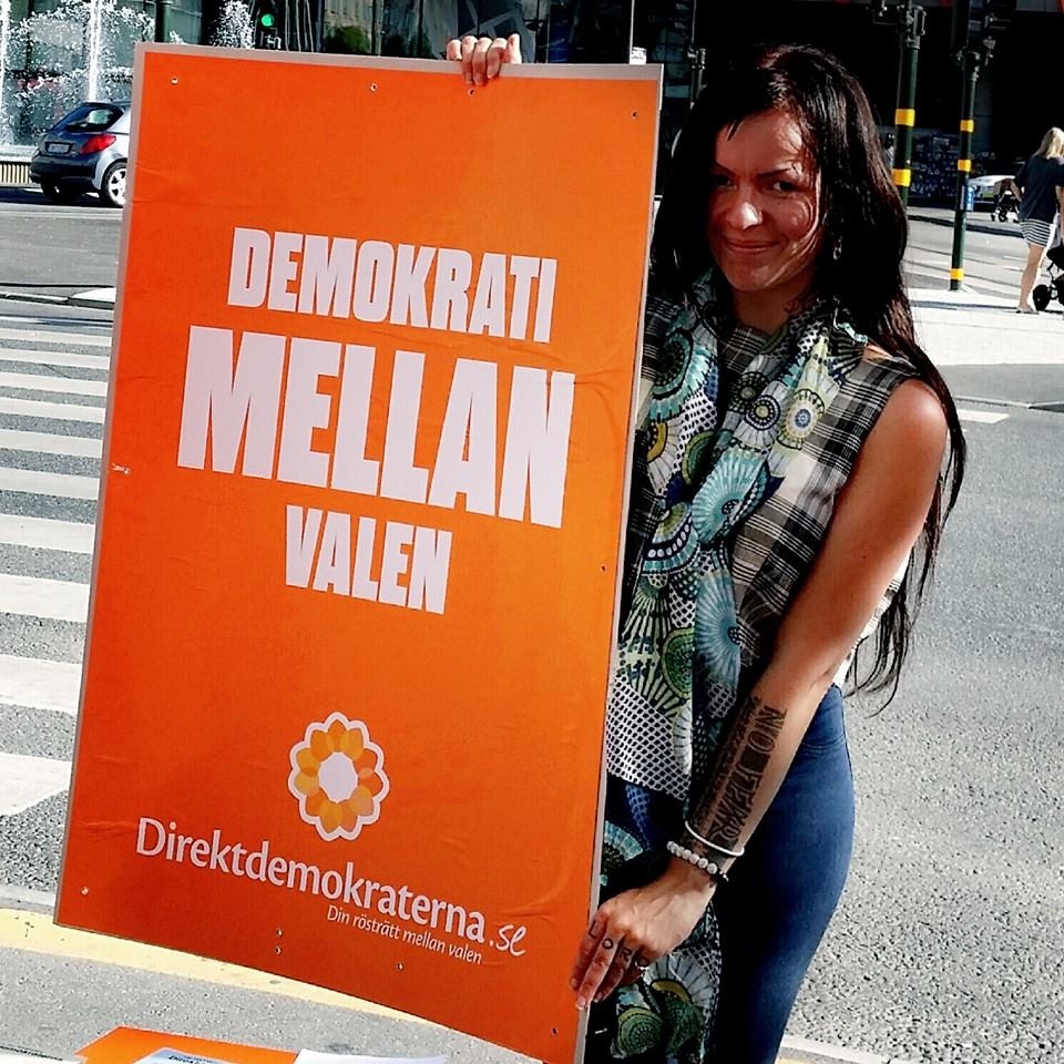 Medlem håller upp en orange kampanjskylt med texten Demokrati mellan valen på en gata